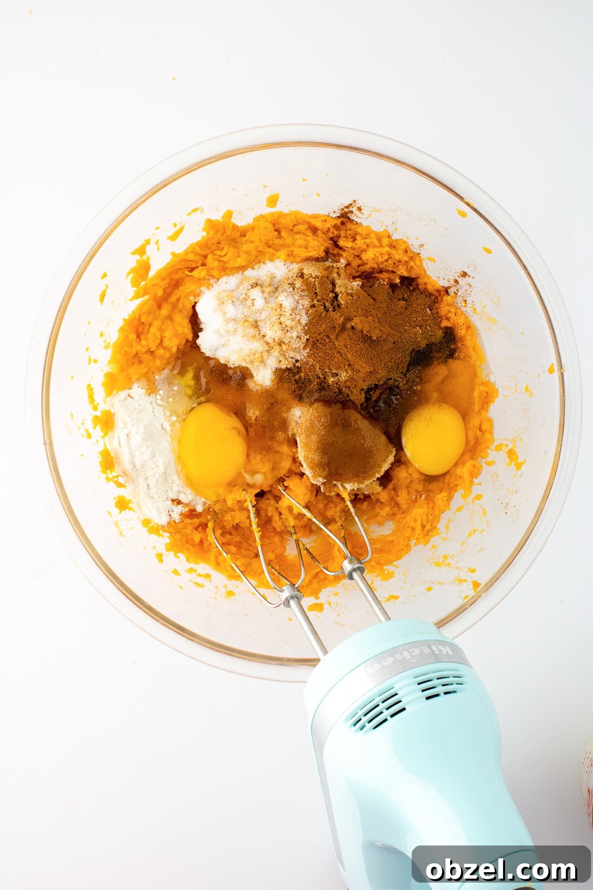 A glass bowl with pumpkin puree, eggs, sugar, flour, spices, and browned butter is being mixed with a light blue electric hand mixer on a white surface—perfect for making a luscious sweet potato pie.