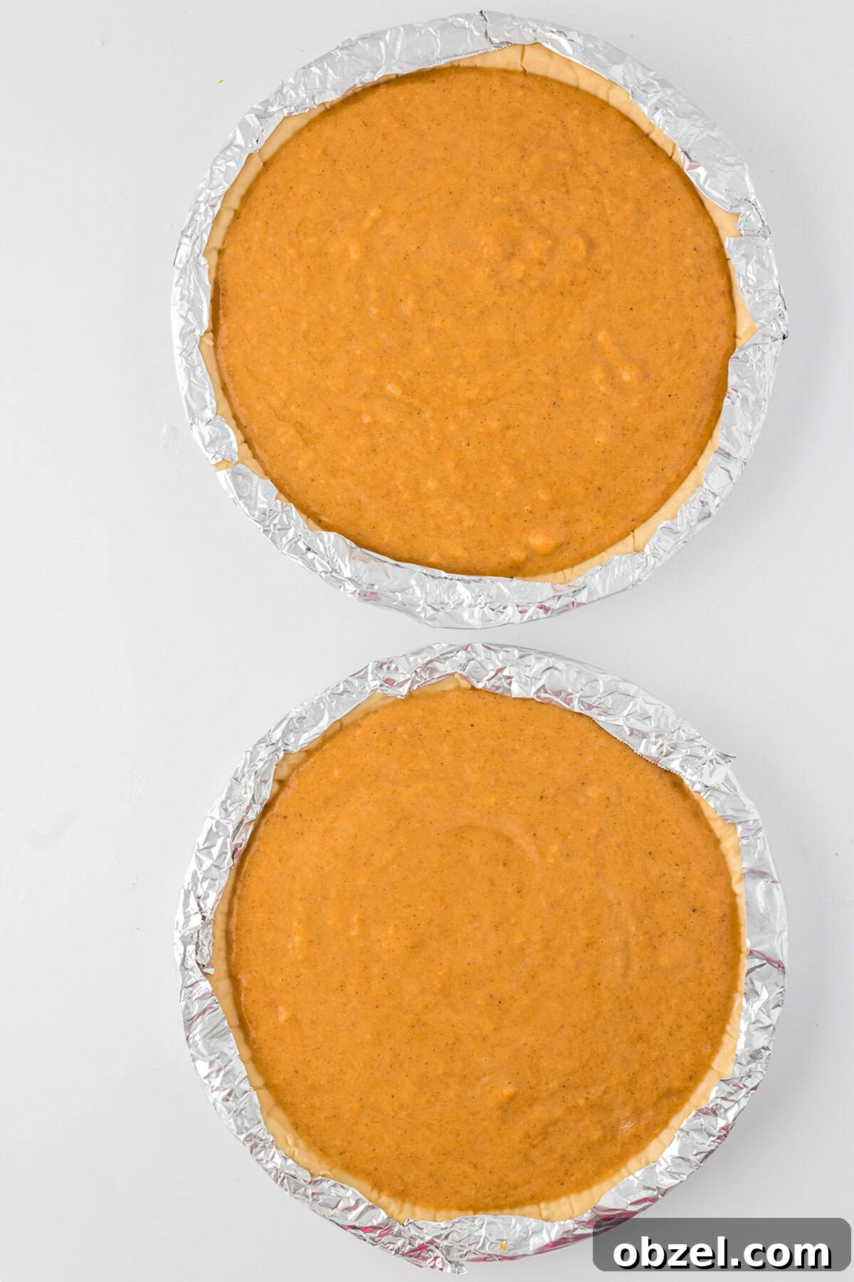 Two unbaked pies—pumpkin and sweet potato—nestle in foil-wrapped tins on a white surface, viewed from above. Imagine them enhanced with the nutty aroma of browned butter for an extra layer of flavor.