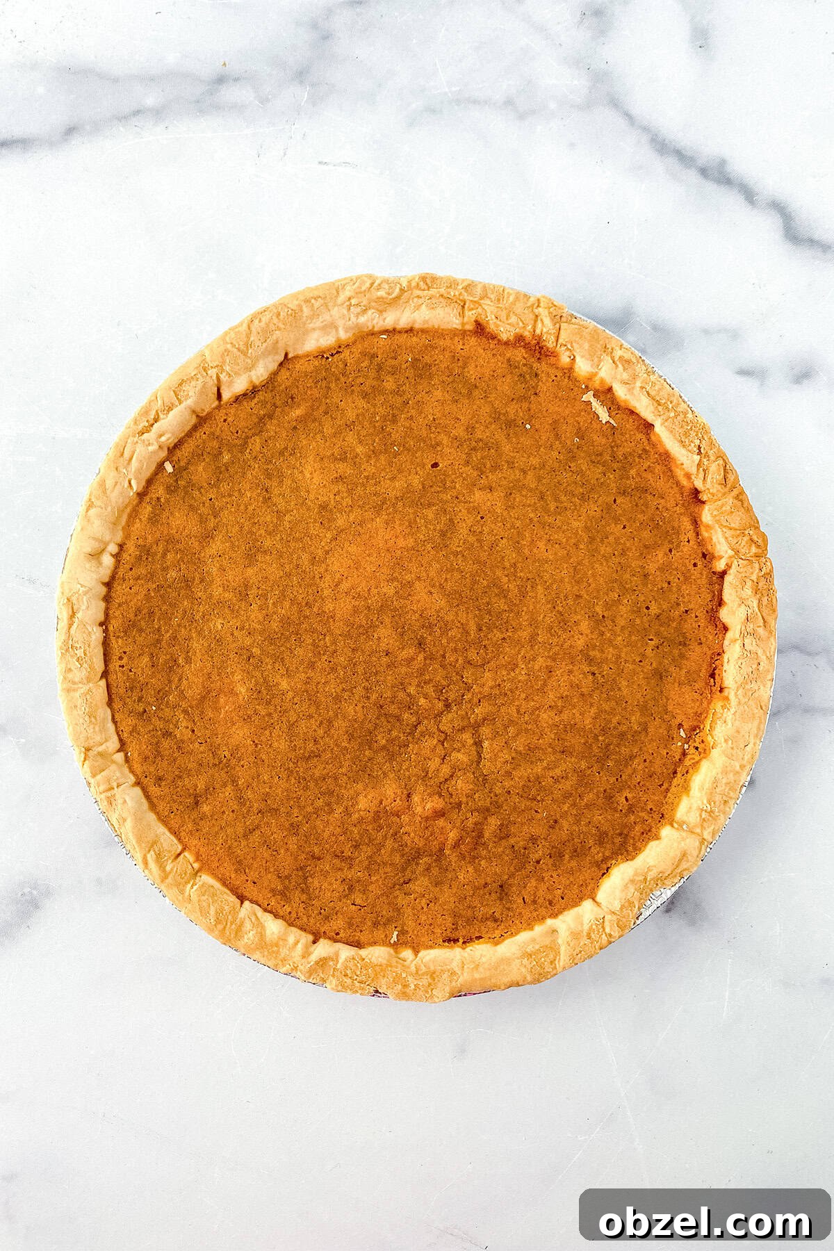 A whole sweet potato pie with a golden-brown filling and crust, featuring hints of browned butter, photographed from above on a white marble surface.