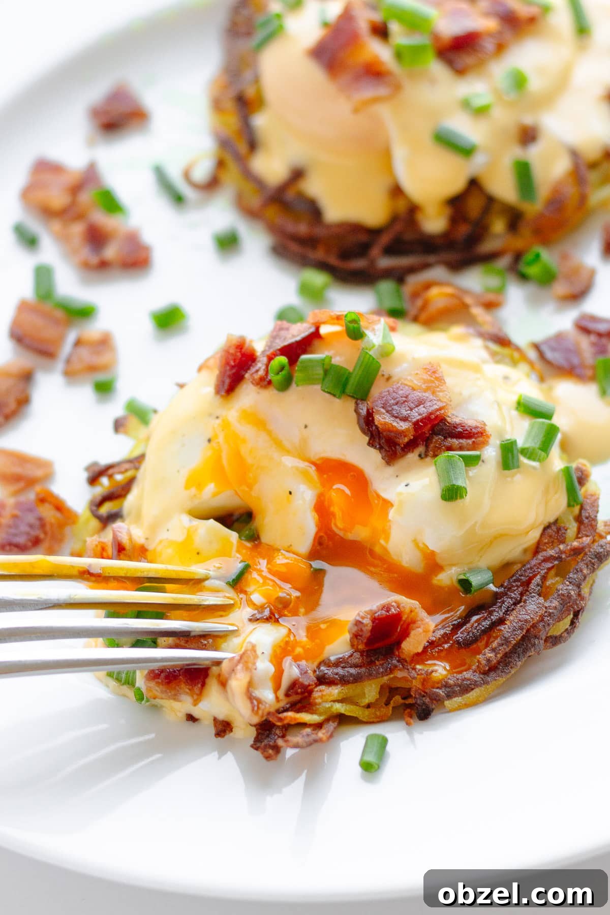Rosti Eggs Benedict with Crispy Potato Pancakes 2 Potato rosti eggs Benedict garnished with chopped chives and crispy bacon pieces with a fork cutting into one.