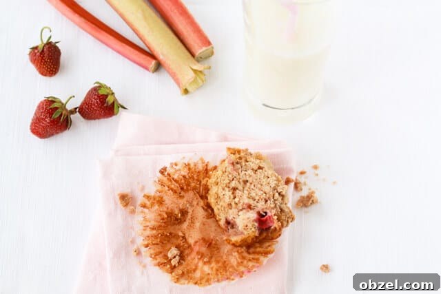 Half of a strawberry rhubarb muffins with crumble topping on a pink napkin with a glass of milk, and a few strawberries and stalks of rhubarb displayed nearby