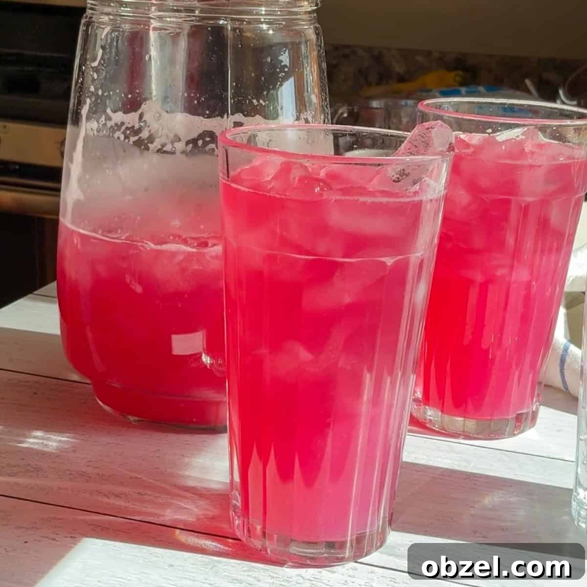 Two glasses of refreshing blueberry lemonade with a pitcher of more to be poured.